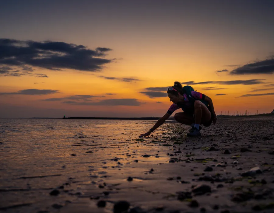 Woman knelt down on sandy beach with sunset behind her