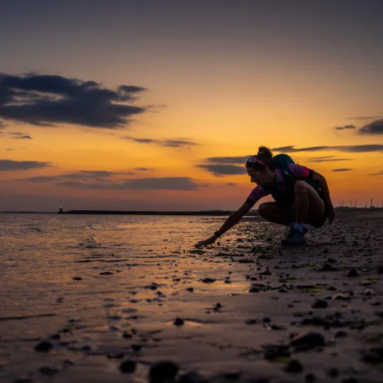 Woman knelt down on sandy beach with sunset behind her