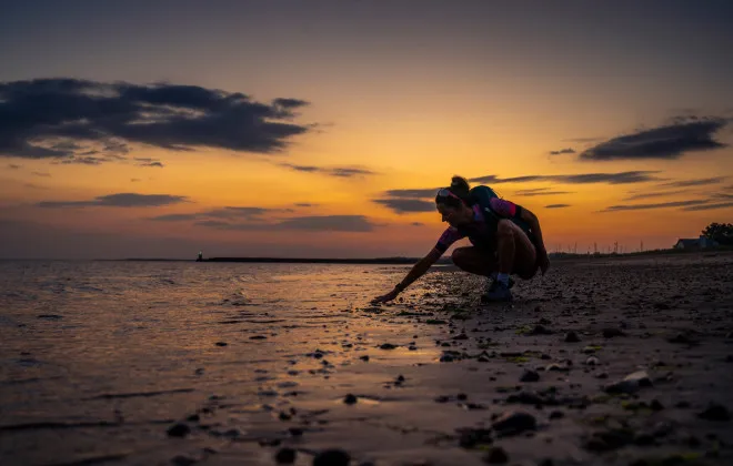 Woman knelt down on sandy beach with sunset behind her